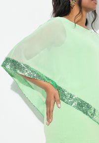 Top verde chiaro con maniche a cappuccio trasparenti, caratterizzato da un orlo decorato con scintillante bordo in paillettes verdi. Tessuto liscio con un'elegante caduta.