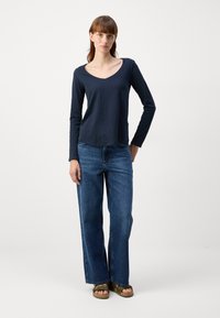 Navy long-sleeve top with a V-neck, paired with high-waisted, wide-leg blue jeans and brown sandals, standing against a plain background.