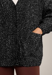 Knitted cardigan in black with white specks. Features a v-neck, button closure, and side pockets. Ribbed cuffs and hem add texture.