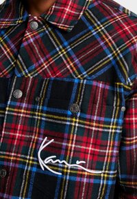 Rutigt skjorta i röd, svart, grön och blå färg. Har metallknappar och en broderad vit logga på framsidan.