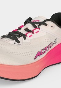 Chaussure de course blanche avec des lacets noirs, des accents roses et le nom de la marque ALTRA sur le côté au-dessus d'une semelle intermédiaire rose et d'une semelle extérieure noire.