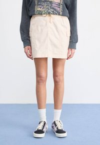 Kort beige skjørt med sidelommer, innsnevret liv, kombinert med svarte og lilla sneakers og hvite ankelsokker. Teksturgrey sweatshirt er brukt.