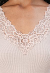 Top ligeiramente rosa claro com textura em nervuras e decote em V, apresentando um intrincado acabamento de renda ao redor do decote, destacando padrões florais e texturas delicadas.