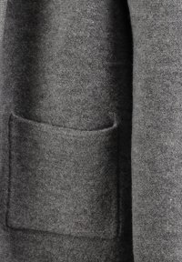 Cappotto grigio in lana con una tasca frontale visibile, texture morbida e linee pulite. Il tessuto presenta un motivo sottile e una finitura opaca.