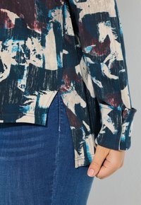 Un top astratto multicolore realizzato in un tessuto morbido, con orlo arrotondato, caratterizzato da uno spacco laterale e maniche lunghe. Abbinato a jeans blu.