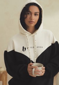 Schwarz-weiß kontrastierende Kapuzenjacke mit Kordelzug und einem Logo auf der Brust, gehalten zusammen mit einer Keramiktasse. Weiche Textur.