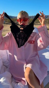 Rövid szőke hajú személy piros napszemüveget visel, és egy fekete csipkebodit tart a kezében, miközben egy csíkos rózsaszín pólóban ül a szabadban.