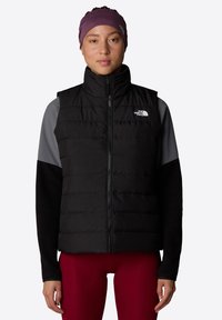 Svart puffervest med glidelås foran, quiltet design, og The North Face-logo. Grå langermede skjorter og tettsittende røde bukser fullfører antrekket.