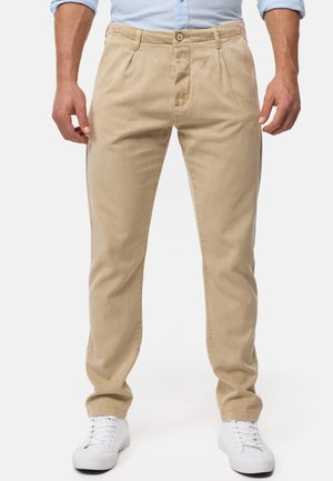 Pantalones de algodón beige con corte recto, con plisados en la parte delantera, cierre de botón y bolsillos laterales, combinados con zapatillas blancas.