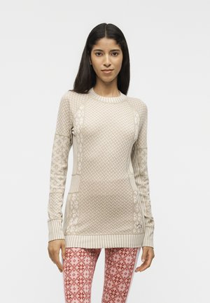 Frau mit langen dunklen Haaren, die einen beigen gemusterten Pullover und rote gemusterte Leggings vor einem einfarbigen weißen Hintergrund trägt.
