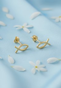 Guldiga fiskformade studs med en slät yta, placerade mot en ljusblå tyg med vita blommopedaler utspridda runtom.