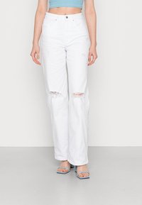 Jeans in denim bianco strappato con ginocchia strappate, vita alta e vestibilità ampia, abbinati a tacchi aperti blu e un crop top coordinato.