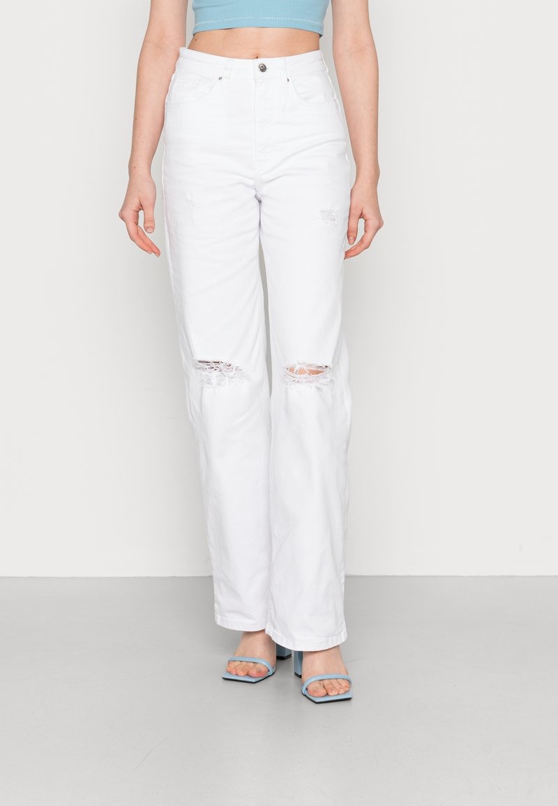 Jeans in denim bianco strappato con ginocchia strappate, vita alta e vestibilità ampia, abbinati a tacchi aperti blu e un crop top coordinato.