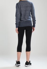 Persona che indossa una maglia a maniche lunghe blu, leggings neri a tre quarti e sneakers nere, in piedi con le spalle rivolte verso di noi su uno sfondo semplice.