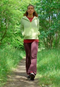 Giacca zip verde chiaro sopra una maglietta rossa, abbinata a pantaloni marrone scuro e scarpe nere, in piedi su un sentiero erboso circondato da alberi.
