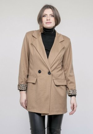 Femme portant un manteau beige à double boutonnage avec des poignets en imprimé léopard, par-dessus un col roulé noir et un pantalon en cuir noir, posant devant un fond uni.