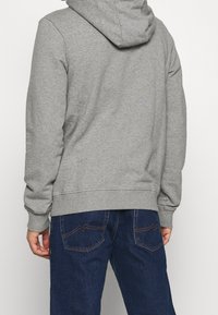 Sudadera gris vista desde atrás, con una textura suave, capucha con cordones y puños y dobladillo acanalados. Combinada con jeans de mezclilla azul.