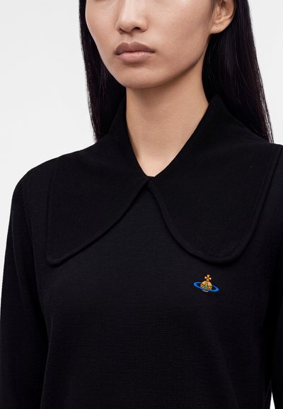 Femme portant un pull noir à large col avec un petit emblème coloré en forme d’orbe sur le côté gauche de la poitrine.