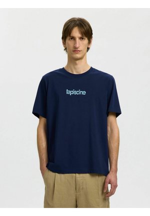 Młody mężczyzna ubrany w granatowy T-shirt z jasnoniebieskim napisem "lapiscine", w połączeniu z beżowymi spodniami, stojący na tle jednolitego jasnego tła.