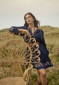Abito in denim ricamato con gonna a campana. Sedia in rattan con accenti a spirale e design circolare. Paesaggio naturale sullo sfondo.