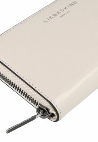 Cartera de color beige claro con exterior de cuero suave, costuras negras y cierre superior con cremallera. Presenta un logo plateado y herrajes duraderos.