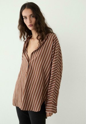 Junge Frau mit langen dunklen lockigen Haaren, die ein lockeres braunes Hemd mit dünnen weißen vertikalen Streifen trägt, steht vor weißem Hintergrund.