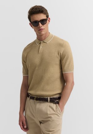 Mann trägt beiges strukturiertes kurzärmeliges Poloshirt, dunkle Sonnenbrille, beige Hose und geflochtenen dunkelbraunen Gürtel, steht mit einer Hand in der Tasche.
