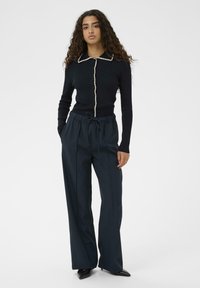 Tânără cu păr lung și creț, purtând un cardigan negru cu nasturi și margini albe, pantaloni bleumarin cu șnur și pantofi negri cu vârf ascuțit.