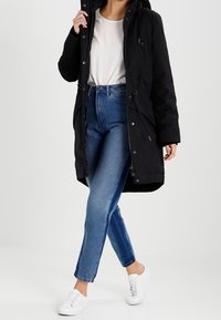 Zwarte parka met een capuchon, met een ritssluiting en een trekkoord in de taille. Draag over een lichtgekleurde top en blauwe jeans, aangevuld met witte sneakers.