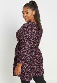 Svart tunika klänning med rosa blommönster; har långa ärmar och en figurnära midja. Materialet verkar lätt och har en slät textur.