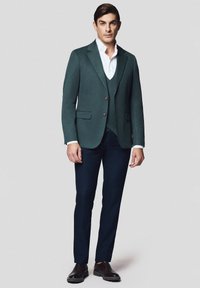 Mężczyzna stojący w zielonej marynarce, dopasowanej kamizelce, białej koszuli, granatowych spodniach i brązowych eleganckich butach na jednolitym tle.