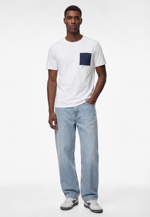 Młody mężczyzna stojący, ubrany w białą koszulkę z granatową kieszenią, jasnoniebieskie dżinsy oraz białe trampki z granatowymi akcentami.