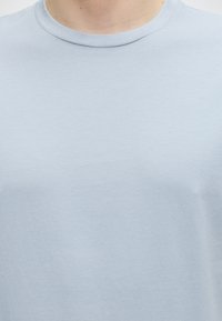 T-shirt bleu clair à col rond porté par une personne, montrant le haut de la poitrine et la zone du cou sur fond blanc.