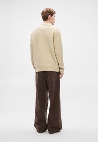 Béžový pletený sveter s vysokým golierom, nosený s voľnými hnedými manžestrovými nohavicami. Výrazné detaily zahŕňajú zaoblený lem a štruktúrovanú látku.