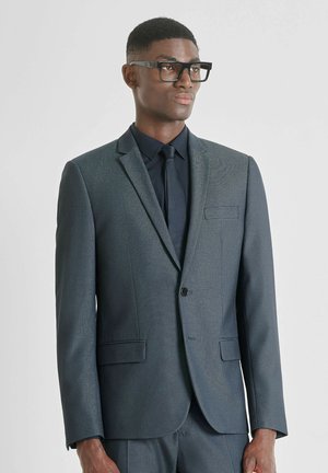 Giacca da suit blu scuro a fantasia con risvolti a punta, chiusura con un solo bottone e due tasche frontali. Indossata sopra una camicia nera e una cravatta.