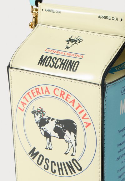 Krems krāsas soma, kas veidota kā piena pakas, ar govs grafiku un "MOSCHINO" birku. Zelta aksesuāri un zila akcents augšpusē.