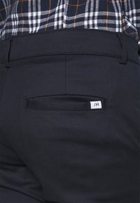 Back pocket of dark navy trousers worn with a navy plaid shirt featuring white and orange lines.