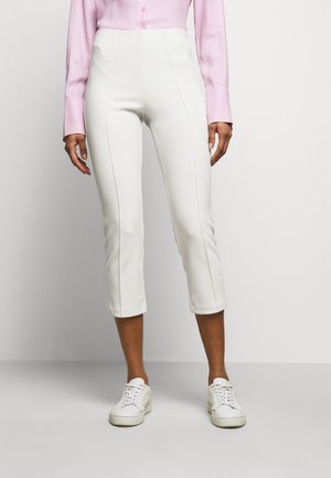 Osoba ubrana w białe, krótkie spodnie, białe trampki i jasnoróżową koszulę na guziki z długim rękawem na jednolitym tle.