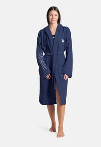 Accappatoio in spugna blu navy con cappuccio, maniche lunghe, cintura in vita e logo "arena" ricamato lungo la manica. Tessuto morbido, lunghezza al ginocchio.