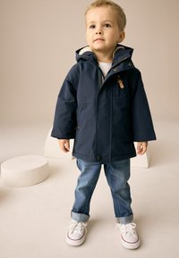Veste imperméable bleu marine avec fermeture éclair et boutons, dotée d'une capuche doublée en fourrure, d'une poche poitrine, portée avec un jean en denim retroussé et des baskets blanches.