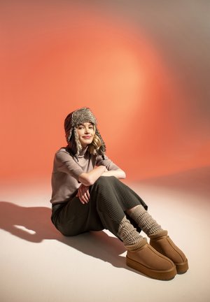 Chapeau en fausse fourrure brun, léger pull, pantalon vert côtelé, et bottines beiges avec des chaussettes en tricot crème épaisses. Style chaleureux et décontracté.