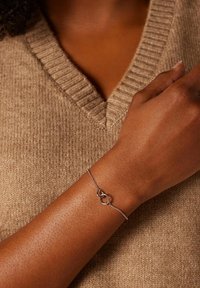 Zilveren schakelschijfarmband met twee in elkaar grijpende ovale schakels. Gemaakt om te dragen om de pols tegen een gestructureerde, beige trui achtergrond.