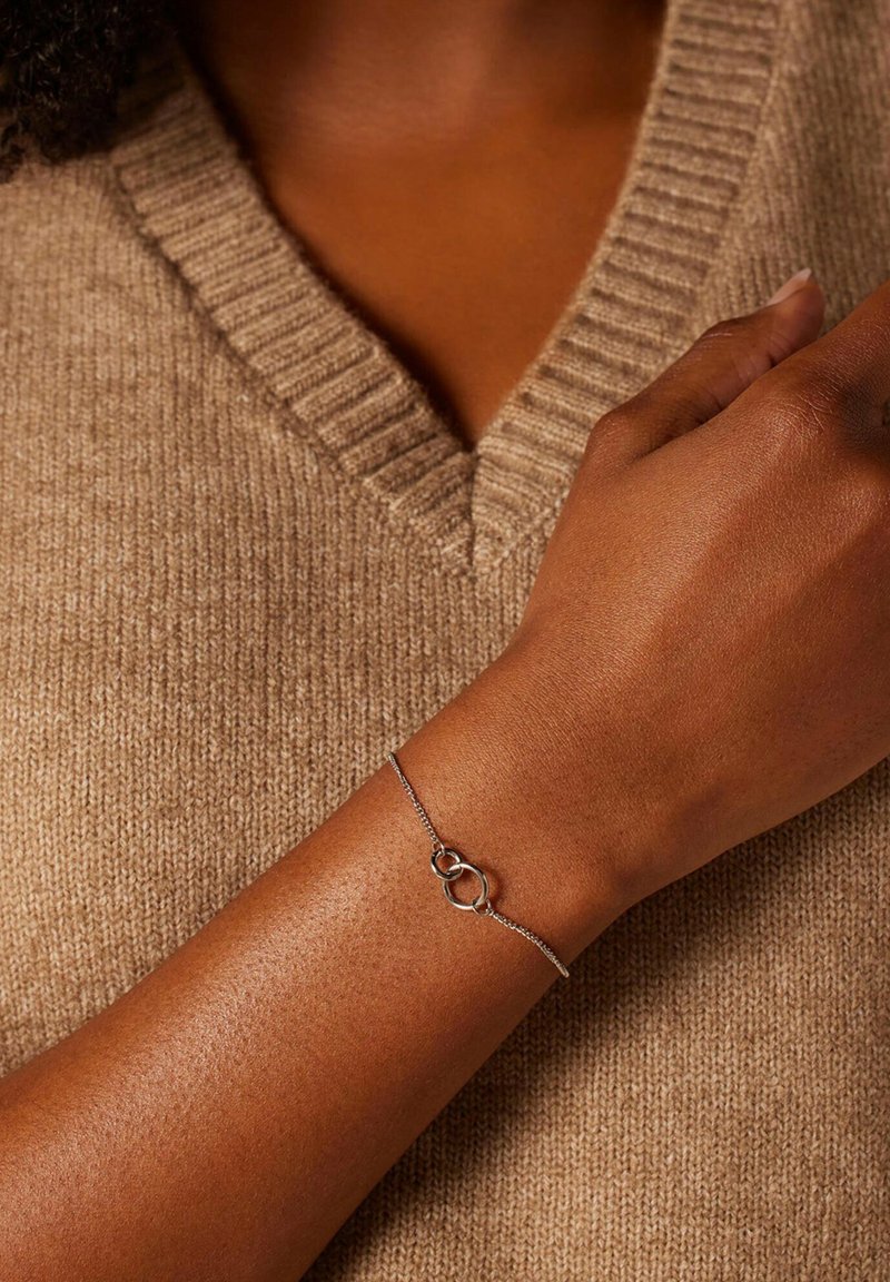 Zilveren schakelschijfarmband met twee in elkaar grijpende ovale schakels. Gemaakt om te dragen om de pols tegen een gestructureerde, beige trui achtergrond.
