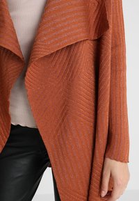 Rust-colored open cardigan with a textured pattern featuring diagonal stripes and a soft, knitted material. Accented with subtle shimmer.