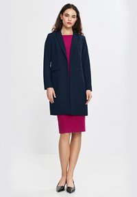 Granatowy długi żakiet z dwoma guzikami i bocznymi kieszeniami, noszony na dopasowanej magentowej sukience. Modelka w czarnych czółenkach, jasne tło.