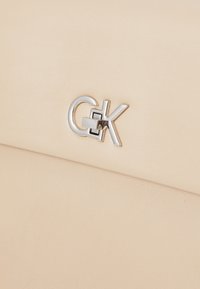 Beige kunstlær clutch med en metallisk logo aksent. Glatt tekstur med subtile korn detaljer, kompakt rektangulær form.