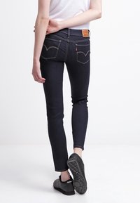 Jeans de mezclilla azul oscuro con un ajuste Slim, bolsillos traseros con una costura curva distintiva y costuras de contraste. Zapatillas negras con suela texturizada.