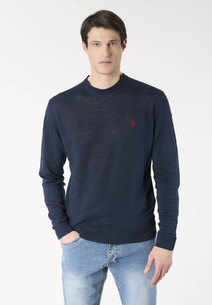 Uomo con capelli scuri che indossa un maglione blu navy con logo rosso sul petto, jeans azzurri, in piedi davanti a uno sfondo bianco uniforme.