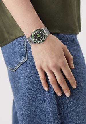 Hand wearing a silver watch with green and black bezel resting on blue denim jeans, next to an olive green shirt hem.
