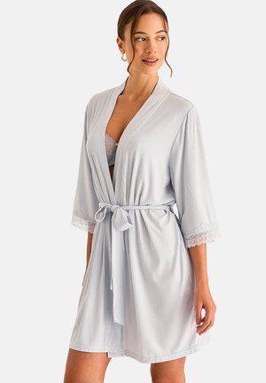 Femme portant un peignoir gris clair mi-genoux avec un galon en dentelle sur les manches, noué à la taille, montrant un soutien-gorge en dentelle gris en dessous, debout sur un fond clair.